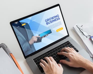 Laptop wyświetlający stronę z nagłówkiem GROWING BUSINESS, na klawiaturze są widoczne ręce osoby.